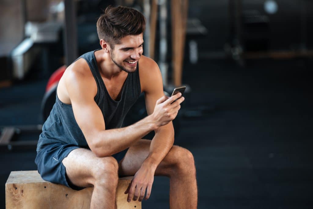 A smiling man sitting on a wooden box in a gym, looking at his smartphone, symbolizing the steps and tools needed to become an online personal trainer.