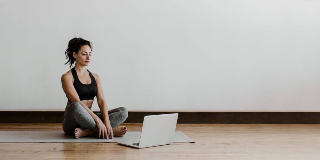 A woman sitting cross-legged on a yoga mat, looking at a laptop in a minimalistic room, symbolizing research and comparison of CEUs for personal trainers.