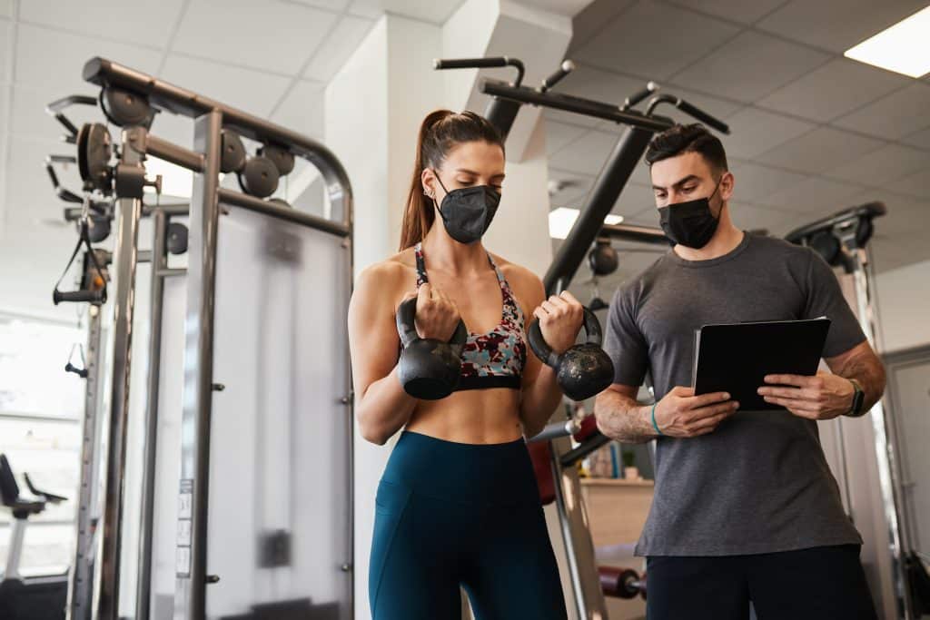 Young fit attractive woman with her personal fitness trainer doing body workout in gym. Keeping social distance and wearing protective face masks. Coronavirus pandemic sport theme.