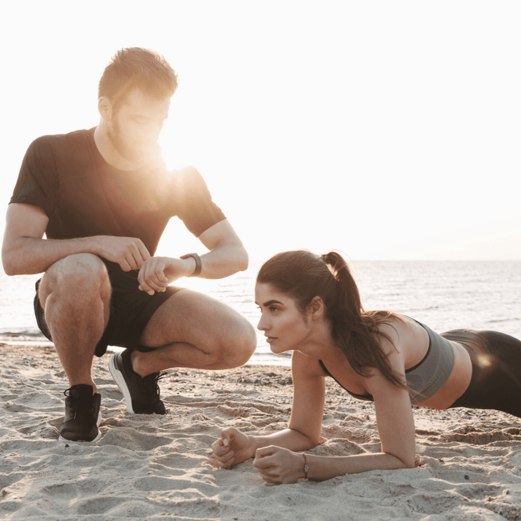 A personal trainer timing a female client as she performs a plank exercise on a sandy beach, depicting a mobile fitness session that could be covered by portable coverage insurance for fitness professionals