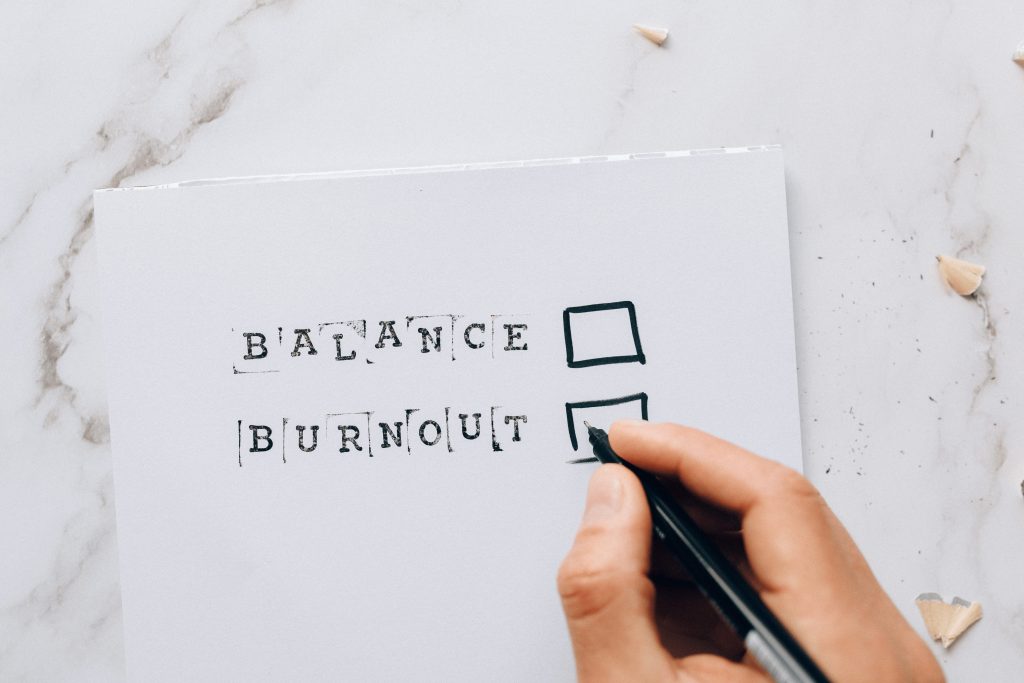 A close-up of a hand holding a black pen poised to mark a checklist. The options 'BALANCE' and 'BURNOUT' are stamped on paper, with 'BURNOUT' unchecked.