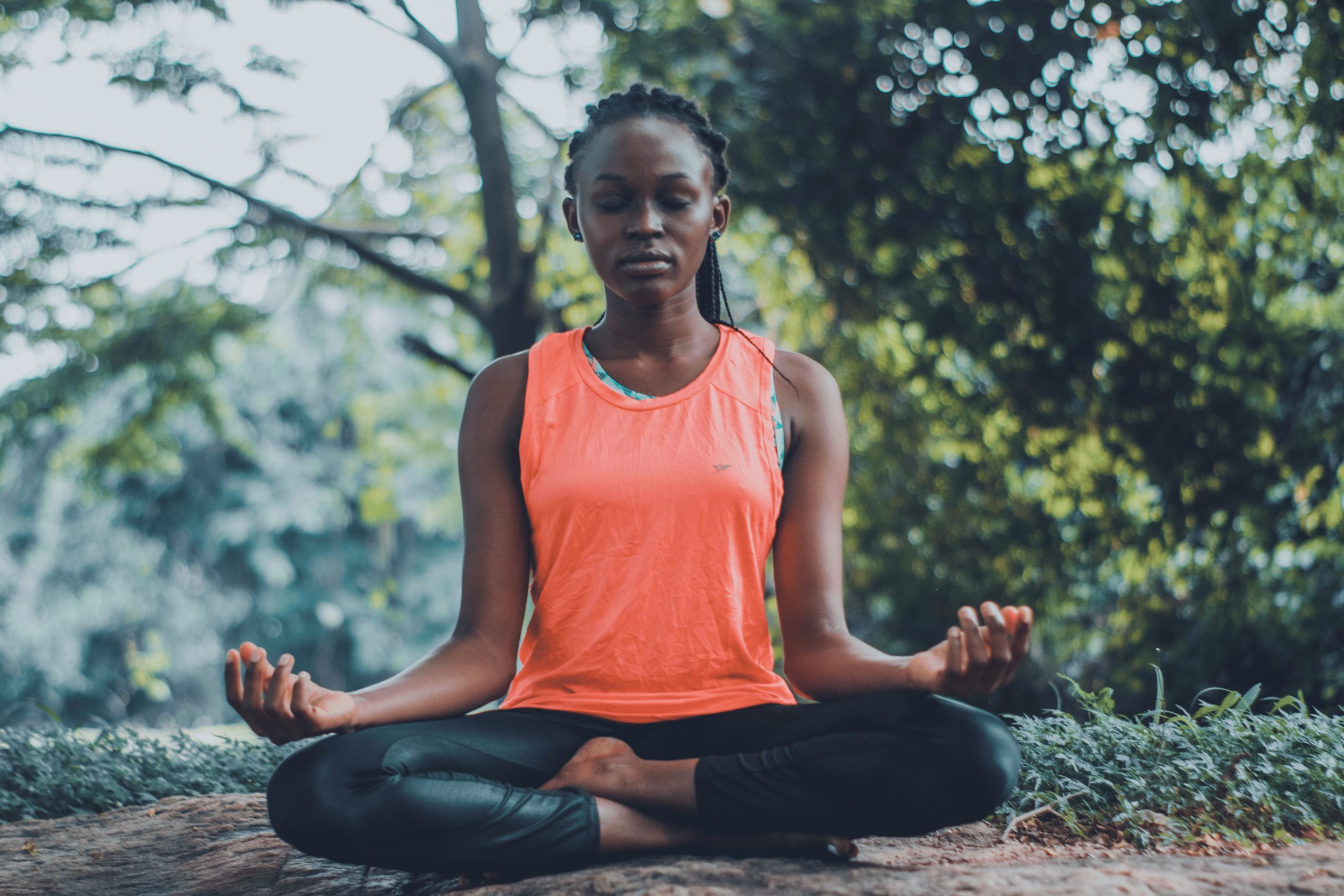 A woman in a vibrant orange tank top sits cross-legged on a large rock outdoors, meditating with closed eyes, surrounded by lush greenery.
