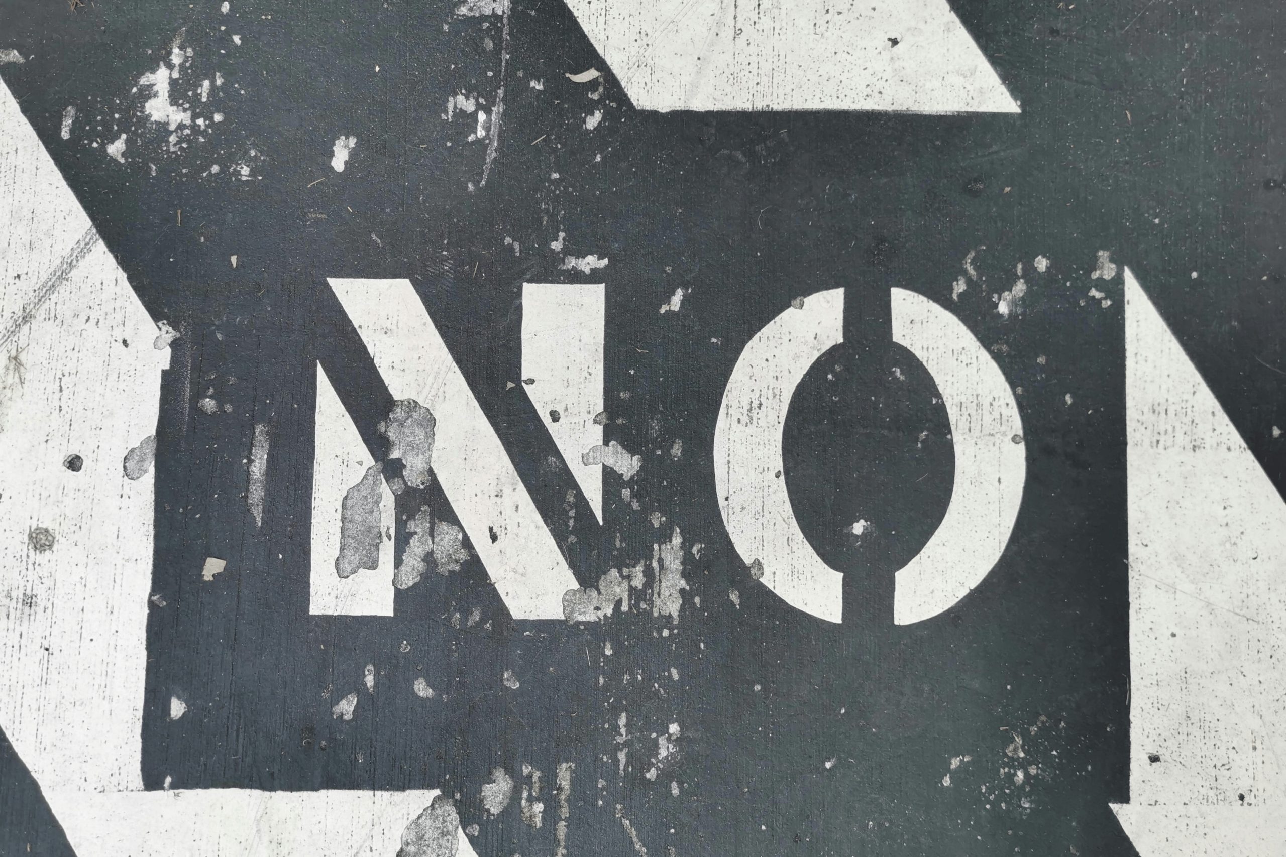 A faded 'NO' text painted on a rough, dark road surface, with signs of wear and peeling paint.