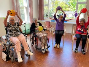 A group of senior women participating in a seated exercise class, holding colorful balls overhead in a bright activity room.