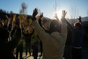 A group of people raising their arms during an outdoor exercise session, with the sun shining in the background and trees in view.