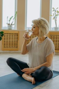A senior woman sitting on a yoga mat, drinking a glass of water after a workout session in a bright indoor space.