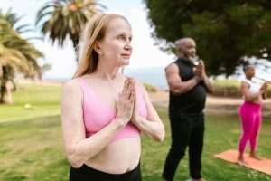 A senior woman practicing yoga outdoors, standing in a prayer pose, with other participants in the background on a sunny day.